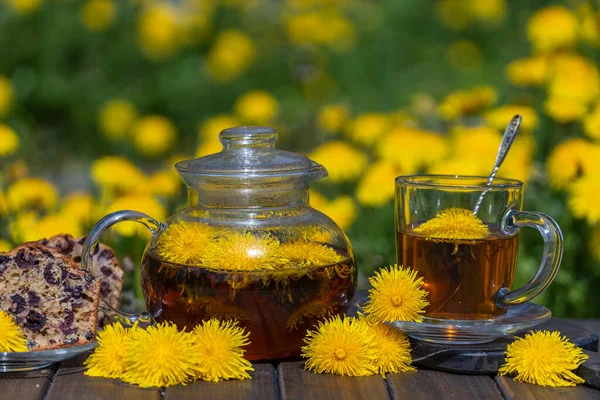 Healthy dandelion flower tea in a glass teapot on the wooden table with cherry muffin in the spring garden, close up